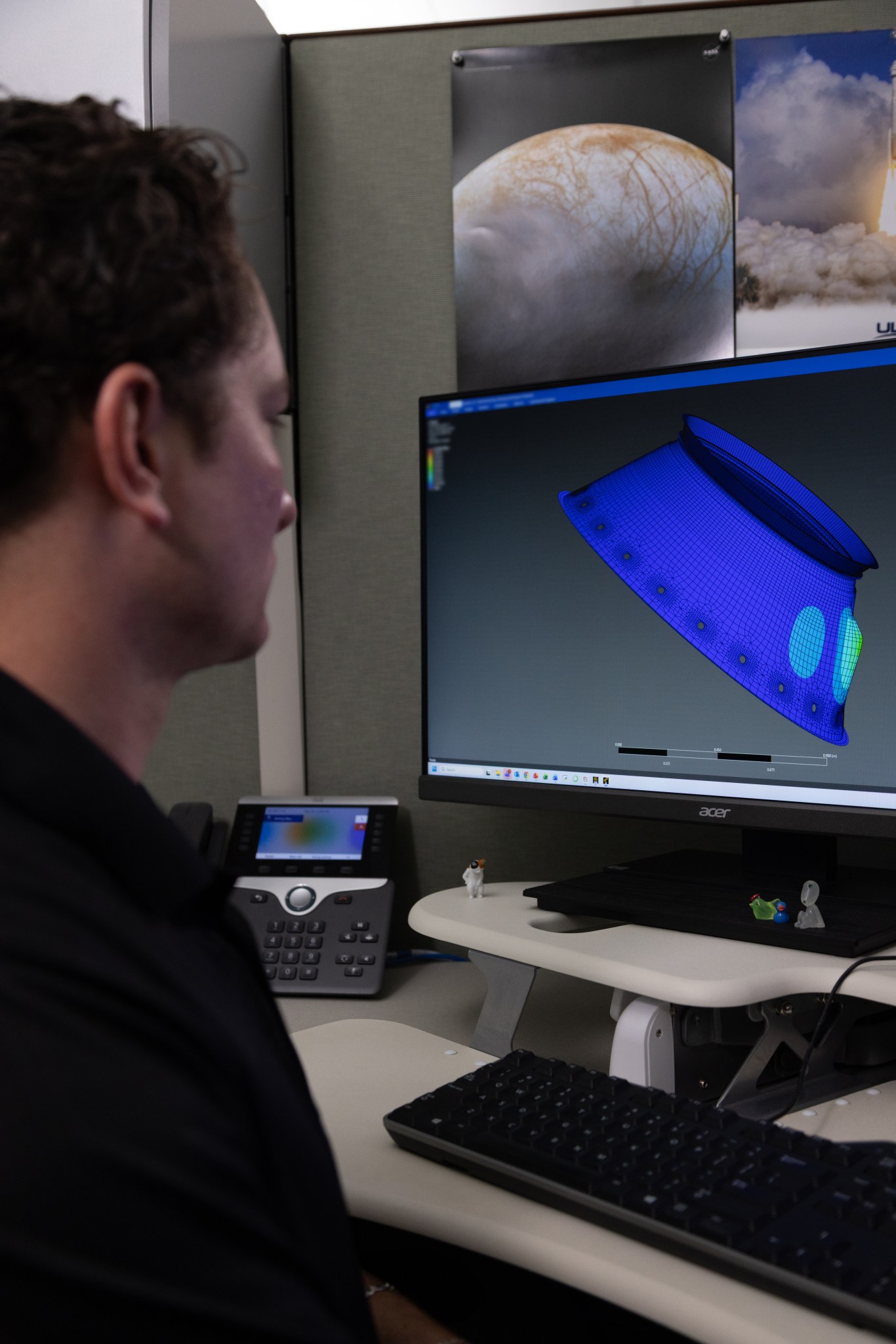 A person sits at a workstation viewing a computer monitor that displays a blue 3D wireframe engineering model of a spacecraft component.