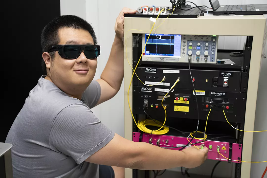 A physicist wearing protective glasses stands beside an electronics rack.